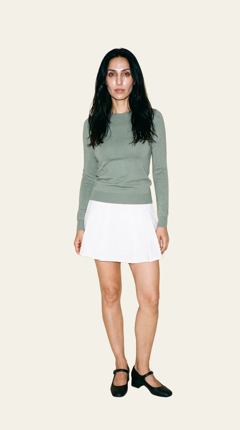 A 42-year-old Arab woman stands in a relaxed pose, shown full body from the front, wearing a white tennis mini skirt. Her confident expression is visible, with a flat alabaster backdrop behind her.