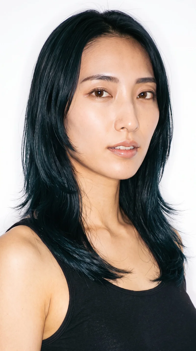 This 28-year-old Japanese woman has a light-tan, warm-yellow undertone and a long face that is more elongated than wide, with a tall forehead and elongated cheeks. Her medium-thickness eyebrows frame her brown eyes, and she has thin lips. Her deep black hair is styled in a layered butterfly cut with shorter face-framing pieces and added volume.