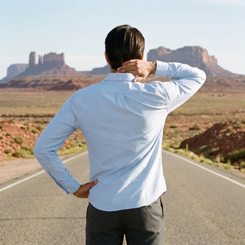 A 20-year-old Southeast Asian male model is shown from the upper body in a back view, wearing a light blue Oxford shirt with one hand behind his neck. A Monument Valley Road backdrop is visible.