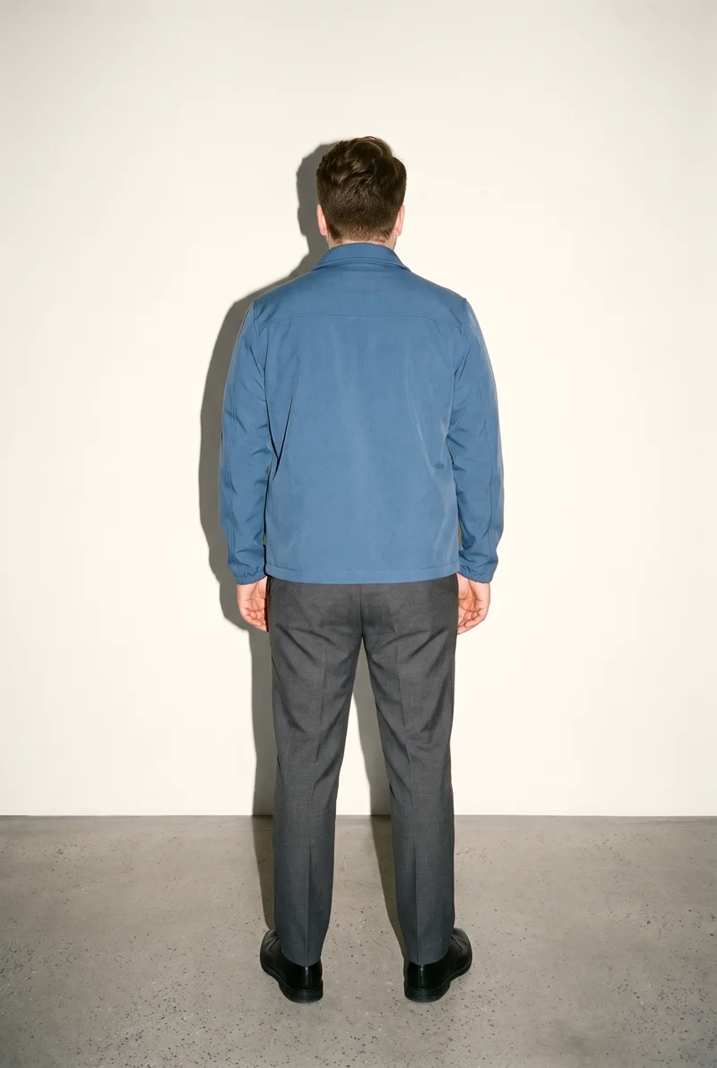 A 29-year-old Caucasian man wears a men's blue softshell jacket in a full-body back view, standing with his ankles crossed. A creamy white studio-wall backdrop frames the composition.