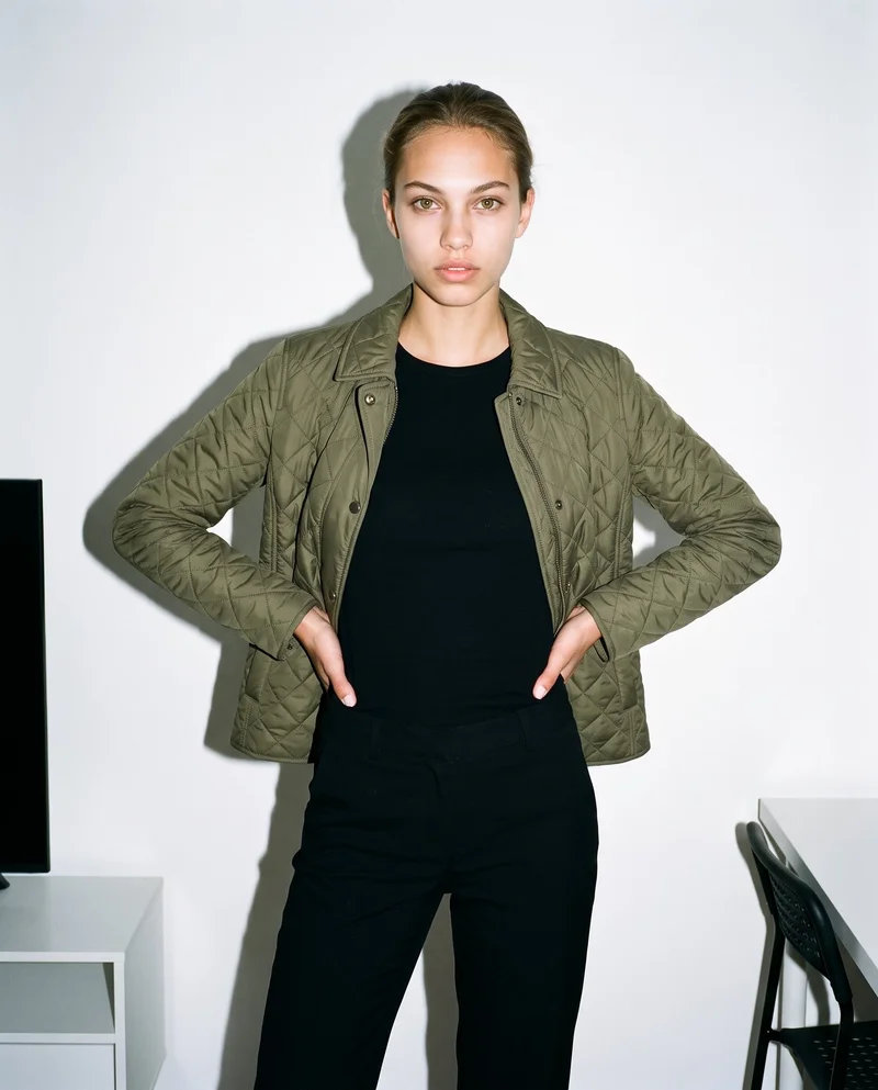 A 19-year-old Caucasian woman wears an olive quilted jacket in a 3/4-length front view, standing with her hands on her hips and a confident expression. A minimalist apartment serves as the backdrop.
