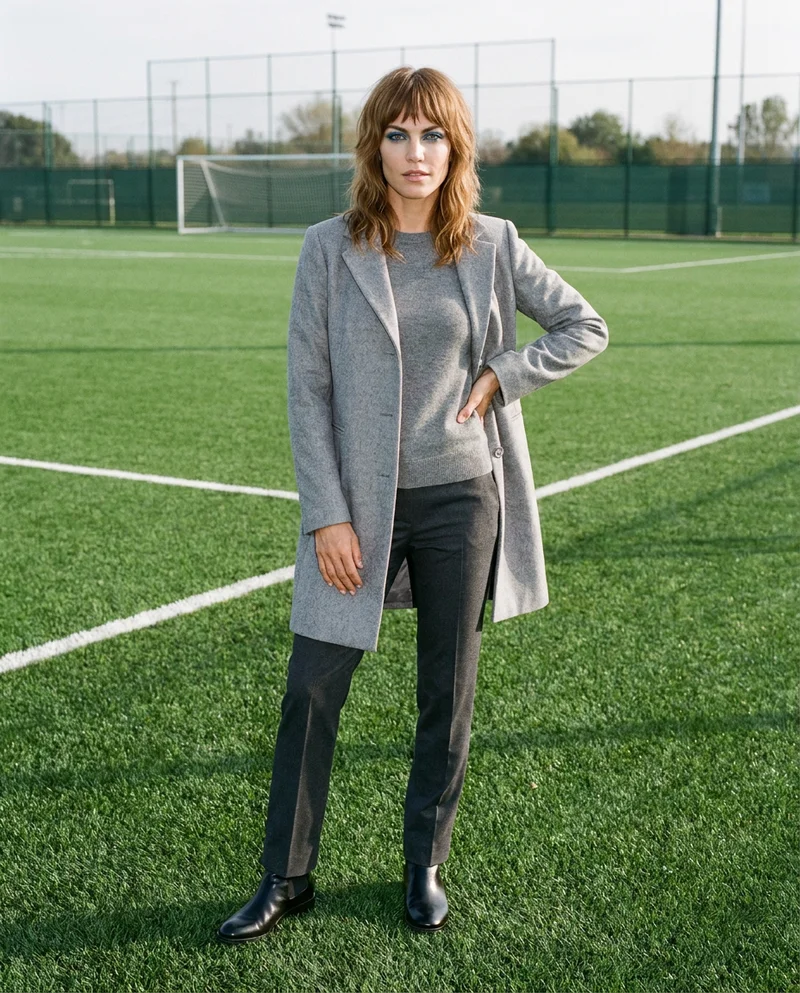 A 36-year-old Caucasian woman wears a women's grey wool coat in a full-body view from the front three-quarter angle. She stands with her weight back and a natural facial expression against a soccer-pitch backdrop.
