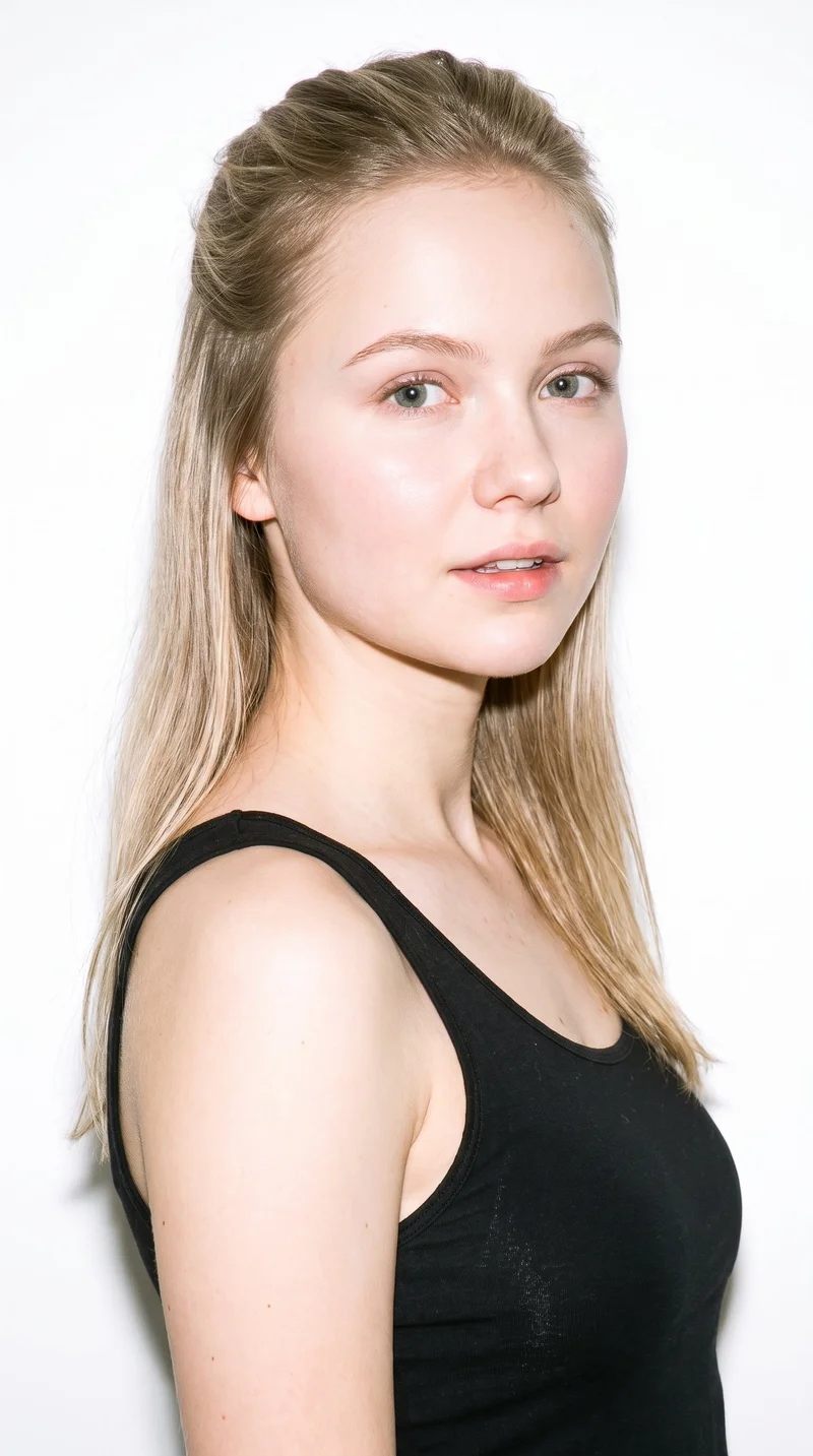 A 20-year-old Slavic woman with fair skin and a cool undertone has a round face with soft, full contours and few angles. Her medium-thickness eyebrows frame her cool, clear grey eyes, and she has thin lips. Her mid-tone blonde hair is styled half up and half down, with the upper section pulled back and the lower lengths hanging loose.