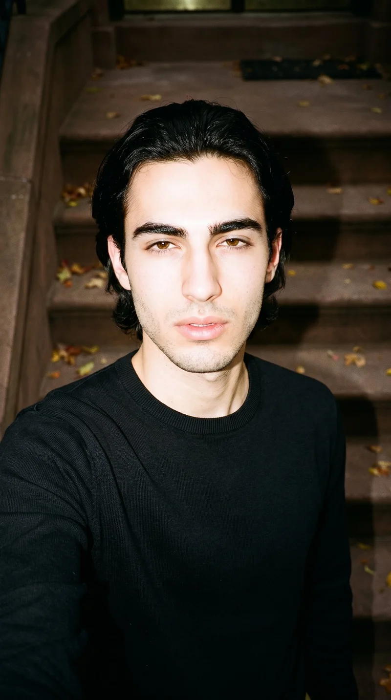 A 19-year-old Caucasian man wears a black pullover jumper in a front-facing selfie with his head level and a sultry expression. A brownstone stoop with autumn leaves forms the backdrop.