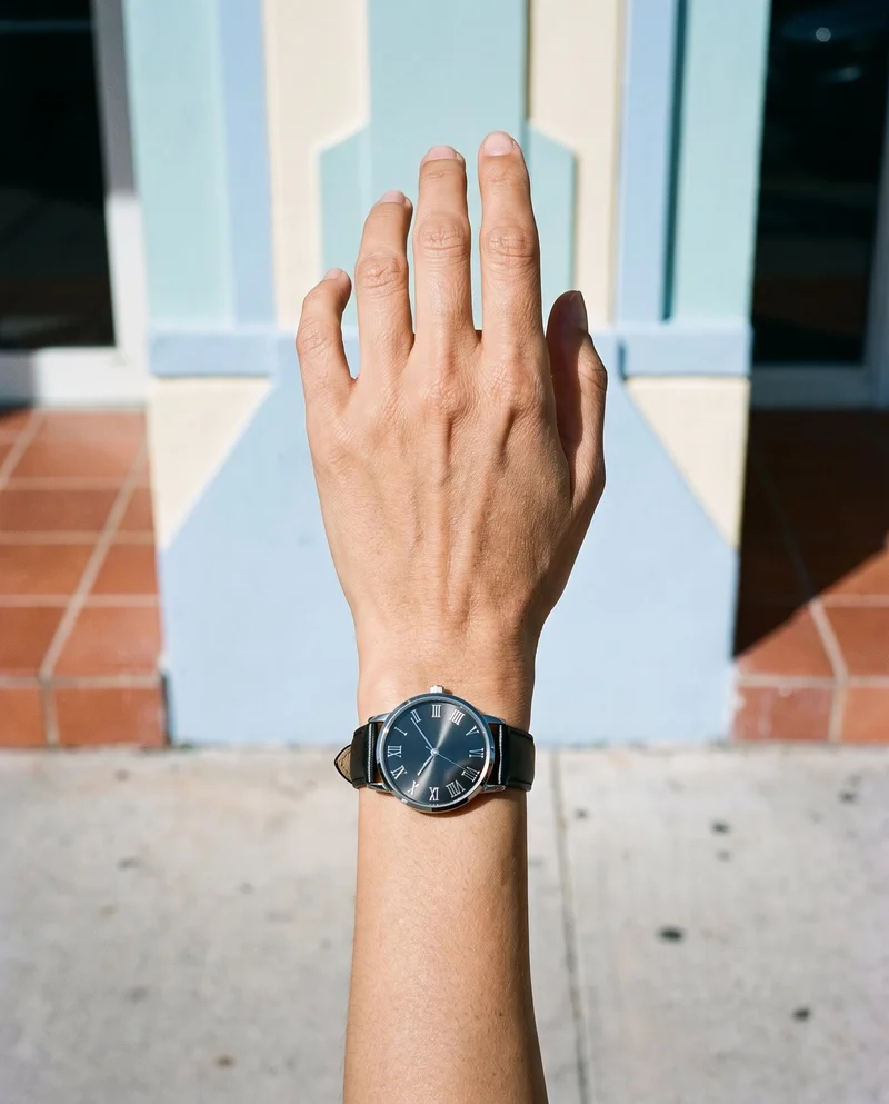 A women's black leather dress watch worn on a relaxed hand and wrist, shown from a top view in a close-up hand-and-wrist frame. The Miami Beach Deco Hotel serves as the backdrop.