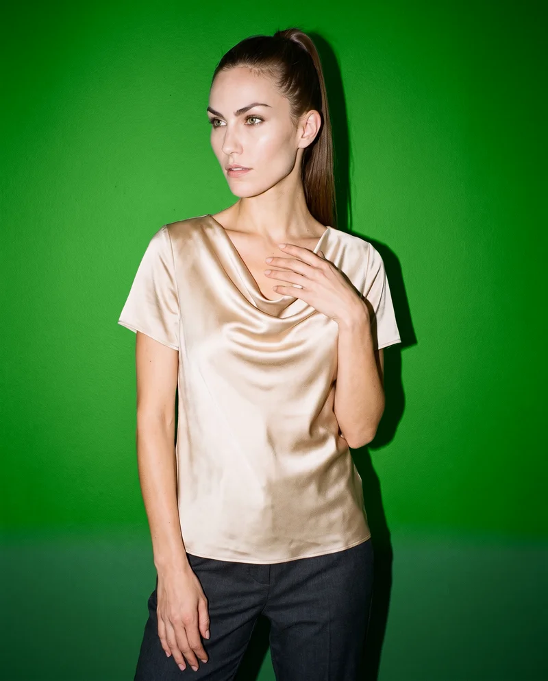 A 28-year-old Caucasian woman wears a champagne satin cowl-neck top, shown from the front at a three-quarter angle in an upper-body frame. Her fingers are held at her chest and her expression is neutral, with a forest-green studio-wall backdrop.