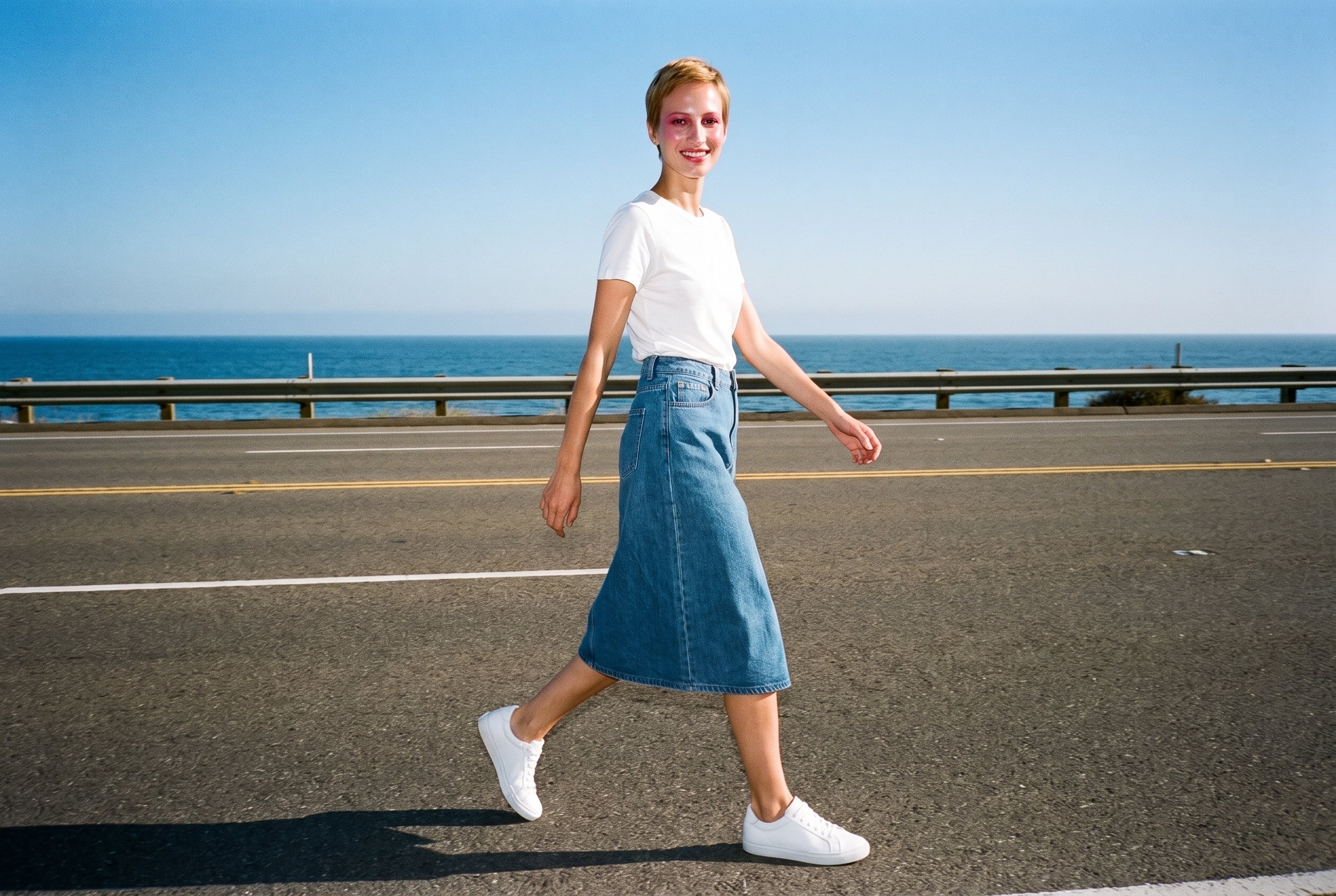 A smiling 25-year-old Caucasian woman is shown full body from an angled front view while walking in a denim A-line skirt. The Pacific Coast Highway appears as the backdrop.