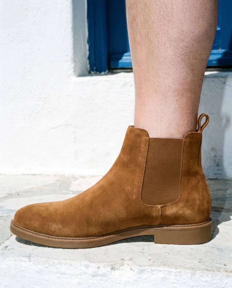 A men's tan suede Chelsea boot shown in an ankle-length side view with the toe pointed. The boot is set against a Greek Villa White Blue backdrop.