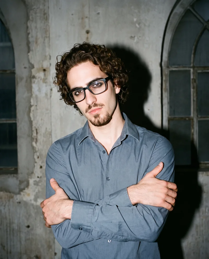 A 26-year-old Caucasian man is shown from the front in an upper-body view, wearing black rectangular optical frames. He holds one elbow and has an intense facial expression. Massive golden loft windows form the backdrop.