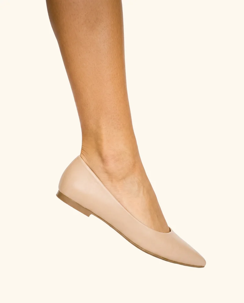 Nude pointed flats shown in a side view with the feet raised on tiptoe, framed from the ankles down against a creamy white backdrop.