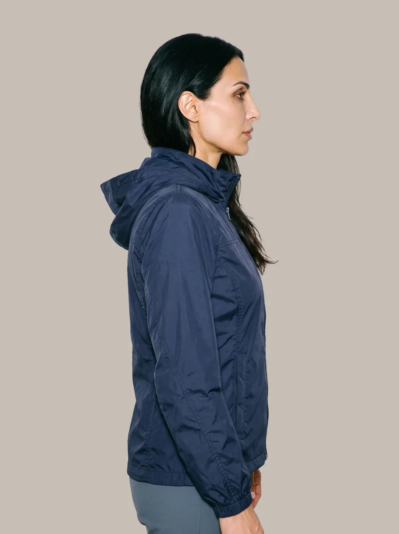 A 42-year-old Arab woman is shown from the upper body in side view wearing a women's navy windbreaker. Her arms are relaxed, and she has a serious facial expression. A flat greige backdrop surrounds her.