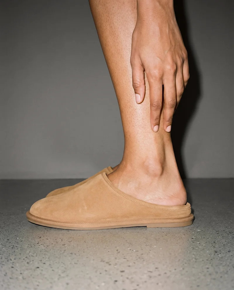 A woman’s ankle and hand are shown in a side view while wearing tan suede slippers. The grey studio wall forms the backdrop.