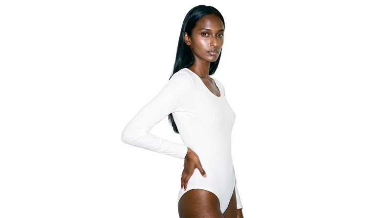 An 18-year-old Indian woman wears a white ribbed bodysuit against a pure white backdrop. She is shown in a three-quarter-length frame from a front three-quarter angle, with one hand on her waist and a natural facial expression.