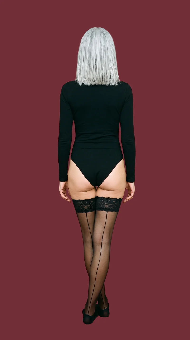 A 42-year-old Arab woman is shown full body from the back wearing black lace hold-up stockings, with her ankles crossed. A flat wine-red backdrop fills the setting.