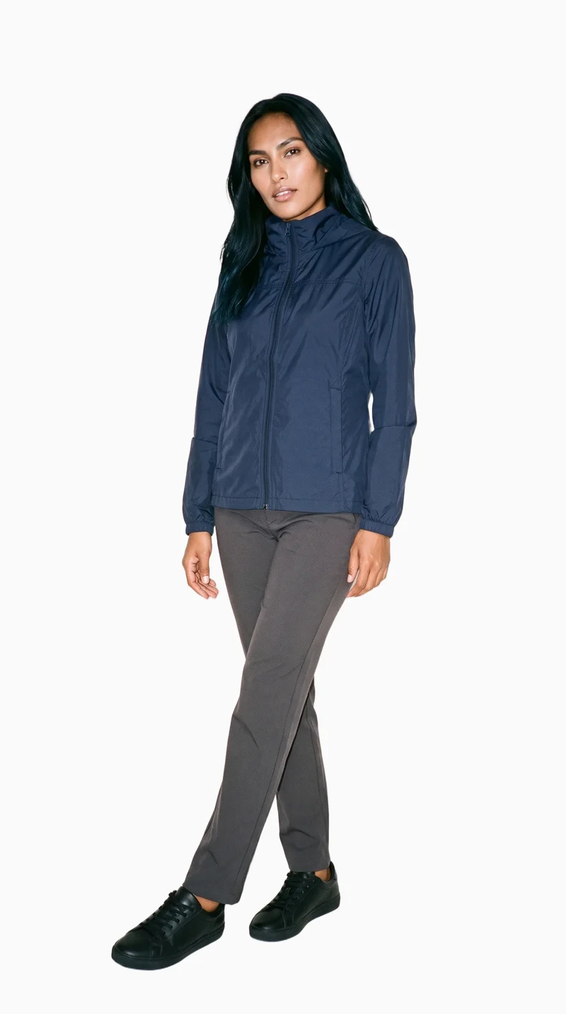 A 24-year-old Indigenous American woman wears a navy windbreaker in a full-body view from a front three-quarter angle. Her ankles are crossed, and the backdrop is a flat Seasalt color.