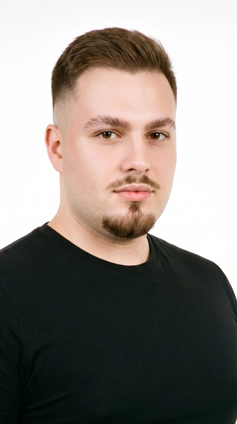 This 28-year-old Caucasian man has medium skin with a cool undertone and a softly structured, refined face with even features. His medium-thickness eyebrows frame brown eyes, and he has full lips. His brown hair tapers from longer on top to very short sides, blending down to the skin. A compact goatee covers his chin while his cheeks remain clear.