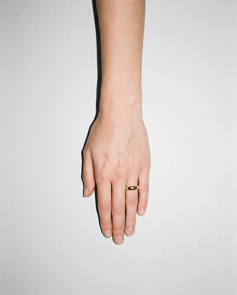 A woman's hand and wrist shown from above, resting flat against a fog grey studio-wall backdrop, with a gold band ring visible on the hand.