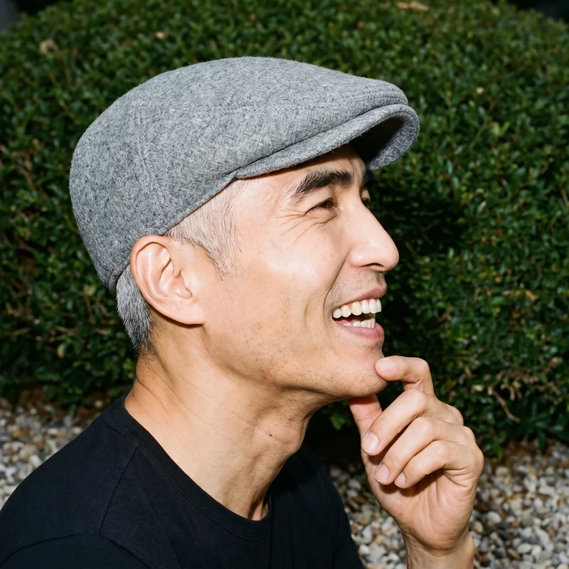 A 54-year-old Korean man is shown in a head-framed side view wearing a grey wool flat cap. He is laughing with one hand at his jaw. A Zen rock garden appears as the backdrop.