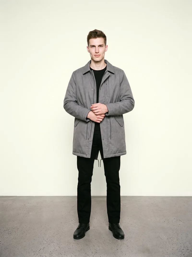 A 21-year-old Caucasian man is shown full body from the front wearing a men's grey longline parka. His hands are held in front, and he has a confident expression. An ivory lace studio backdrop fills the background.
