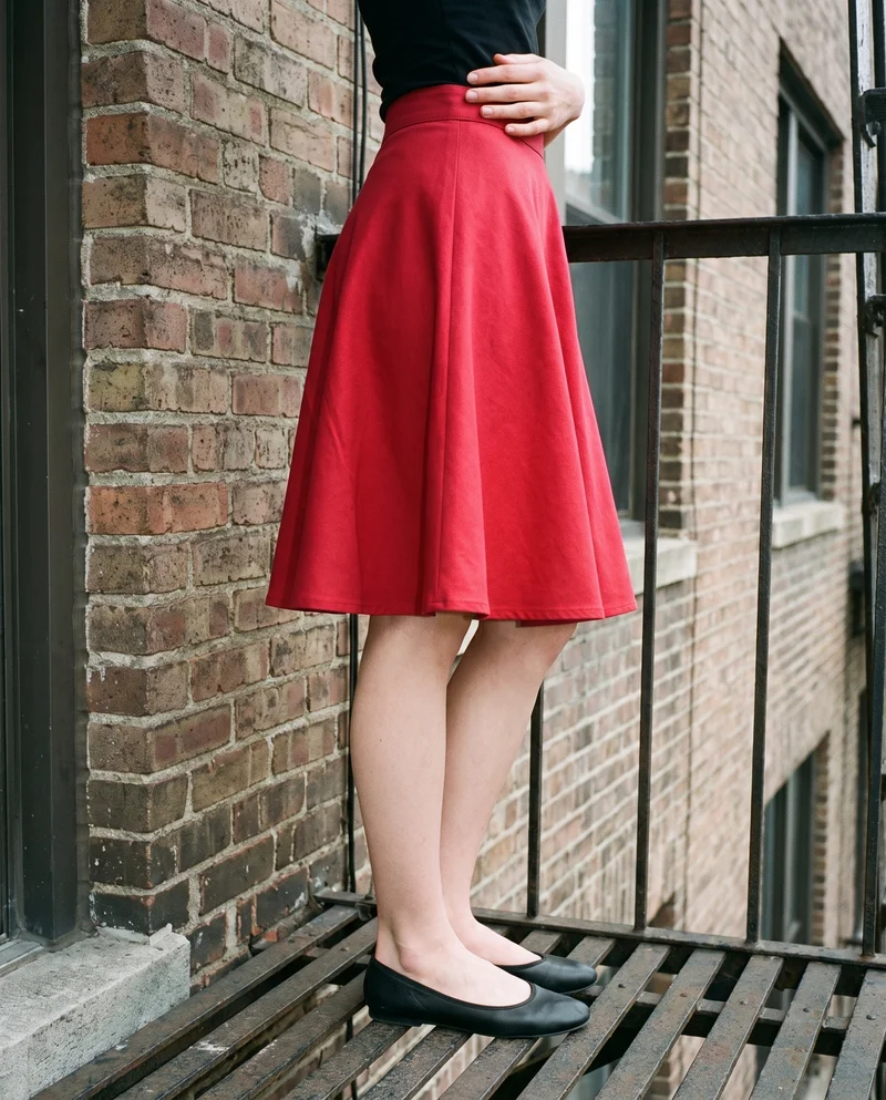A model wears a red A-line skirt, shown from the lower body in side view with her hips turned. A fire escape serves as the backdrop.