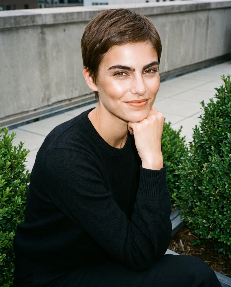 A 25-year-old Caucasian woman wears a black cashmere knit in an upper-body, angled-front view, with her wrist resting at her jaw and a natural expression. The setting is shown against a formal rooftop garden backdrop.