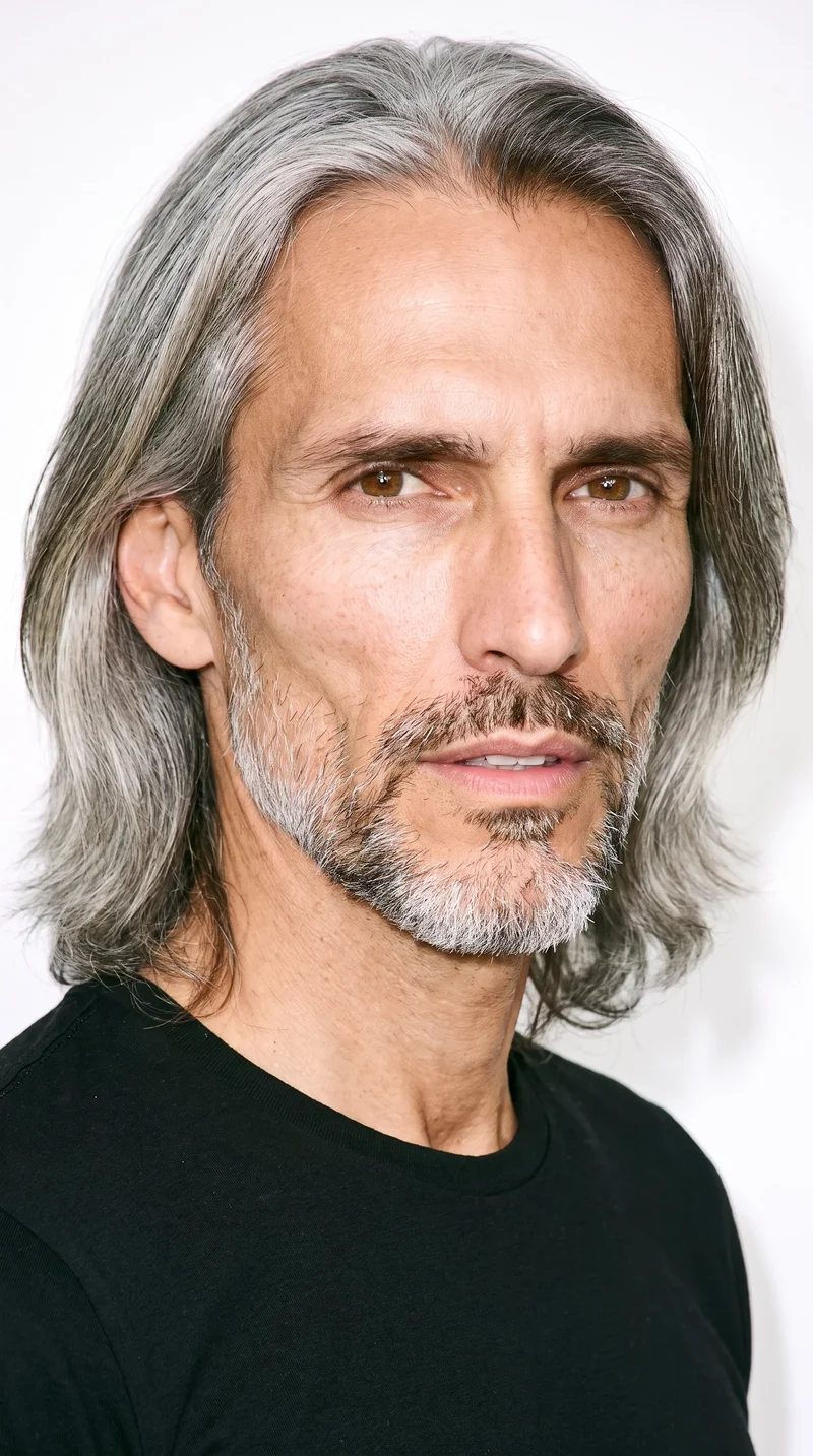 A 47-year-old Hispanic man with medium skin and a neutral undertone has a gaunt face with lean features, hollowed cheeks, and sharply defined bone structure. He has medium-thick eyebrows, brown eyes, and full lips. His gray hair is long, worn loose past the shoulders with natural volume, and he has a short, full beard kept evenly trimmed with clean cheek and neckline edges.