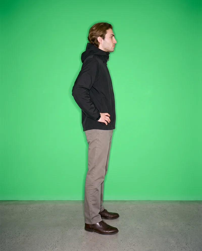 A 23-year-old Caucasian male model wears a black hardshell jacket in a full-body side view, standing in a wide stance with a neutral expression. A dusty emerald studio-wall backdrop fills the background.