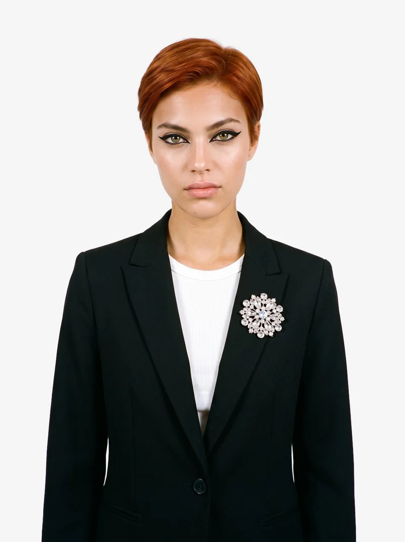 An 18-year-old Caucasian woman is shown from the front in an upper-body view wearing a women's crystal brooch. Her arms are relaxed and her expression is confident, with a flat cloud-white backdrop.