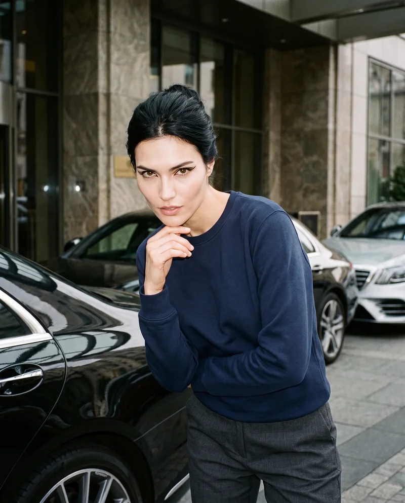 A 23-year-old Hispanic woman wears a navy cotton crewneck sweatshirt in an upper-body front view. She poses with one hand at her chin and a serious expression against a valet stand backdrop.