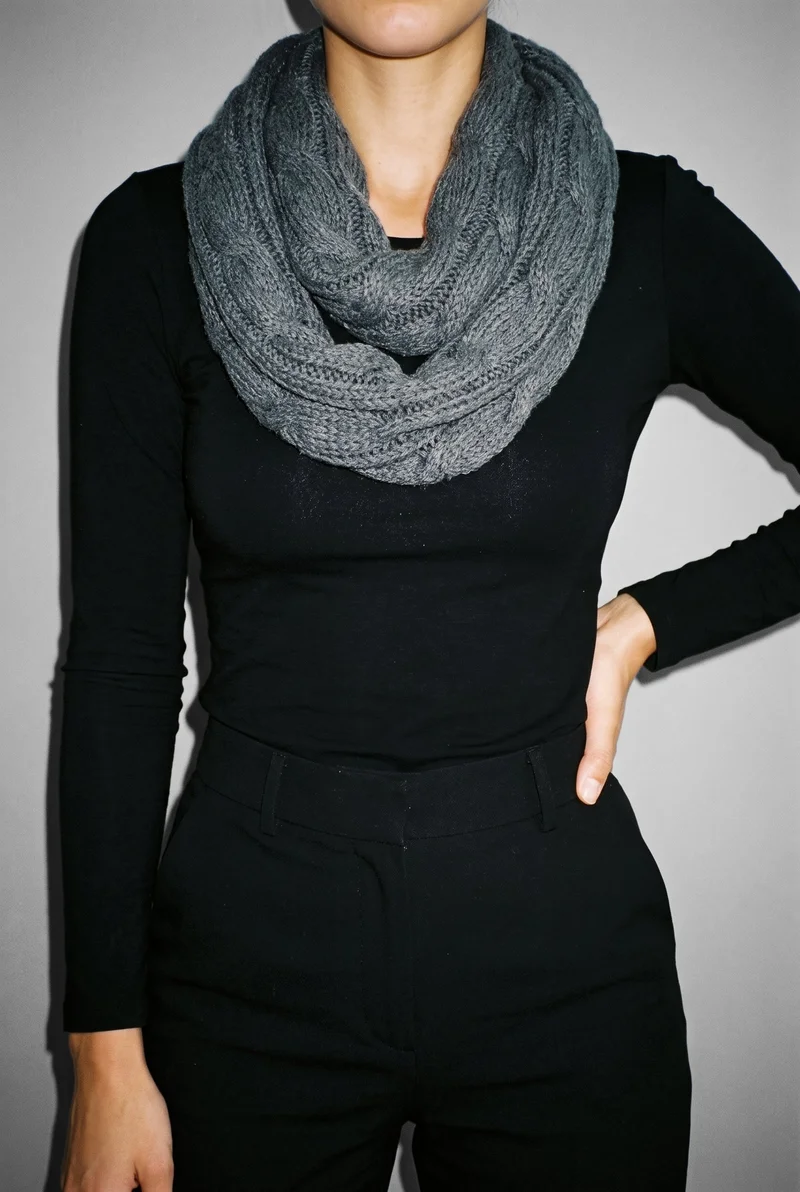 A woman wears a grey knit snood, shown from the front in a torso-length view with one hand on her hip. A pebble grey studio-wall backdrop fills the background.