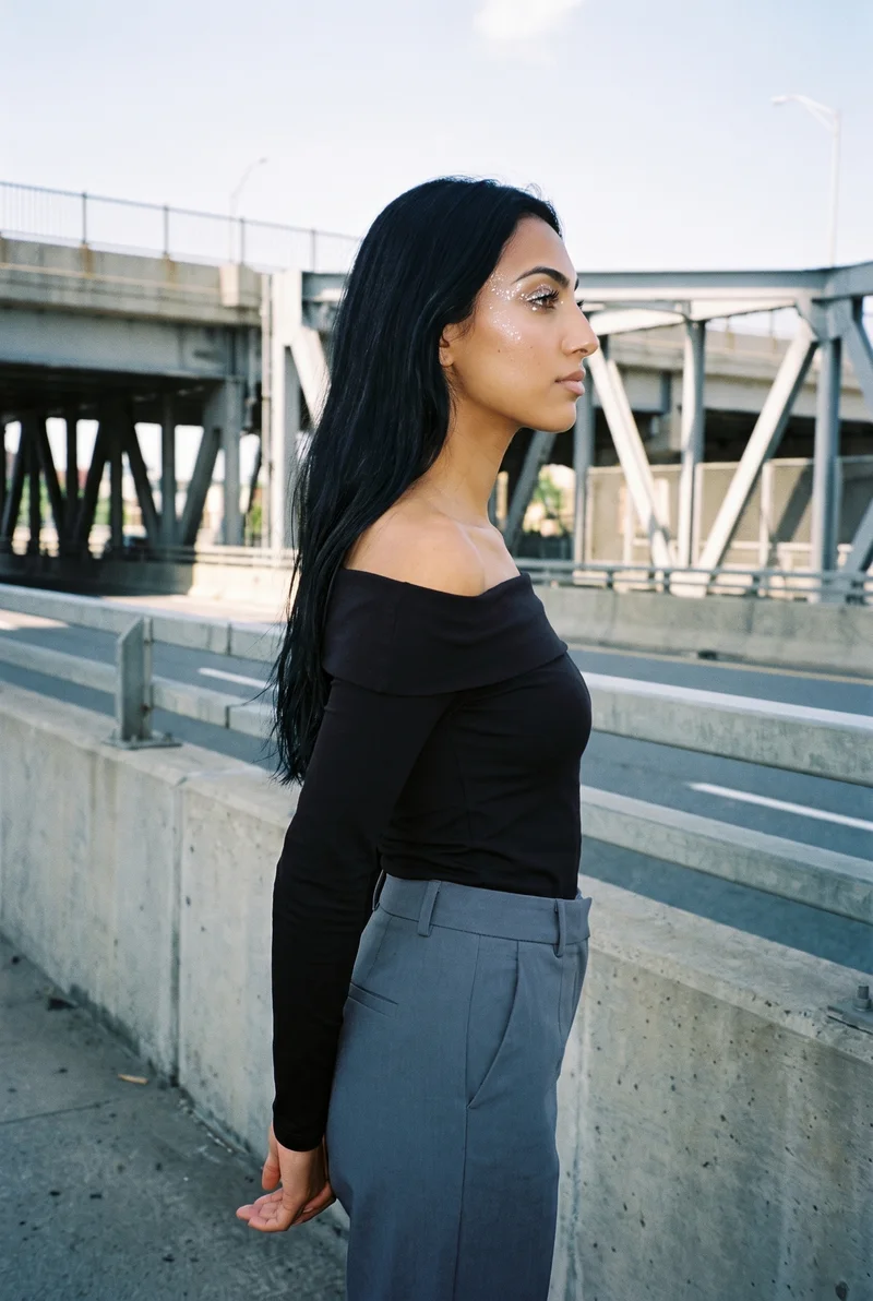 A 19-year-old Arab woman wears a black Bardot top in a three-quarter-length side view, standing with her hands behind her back and a curious expression. A highway overpass forms the backdrop.