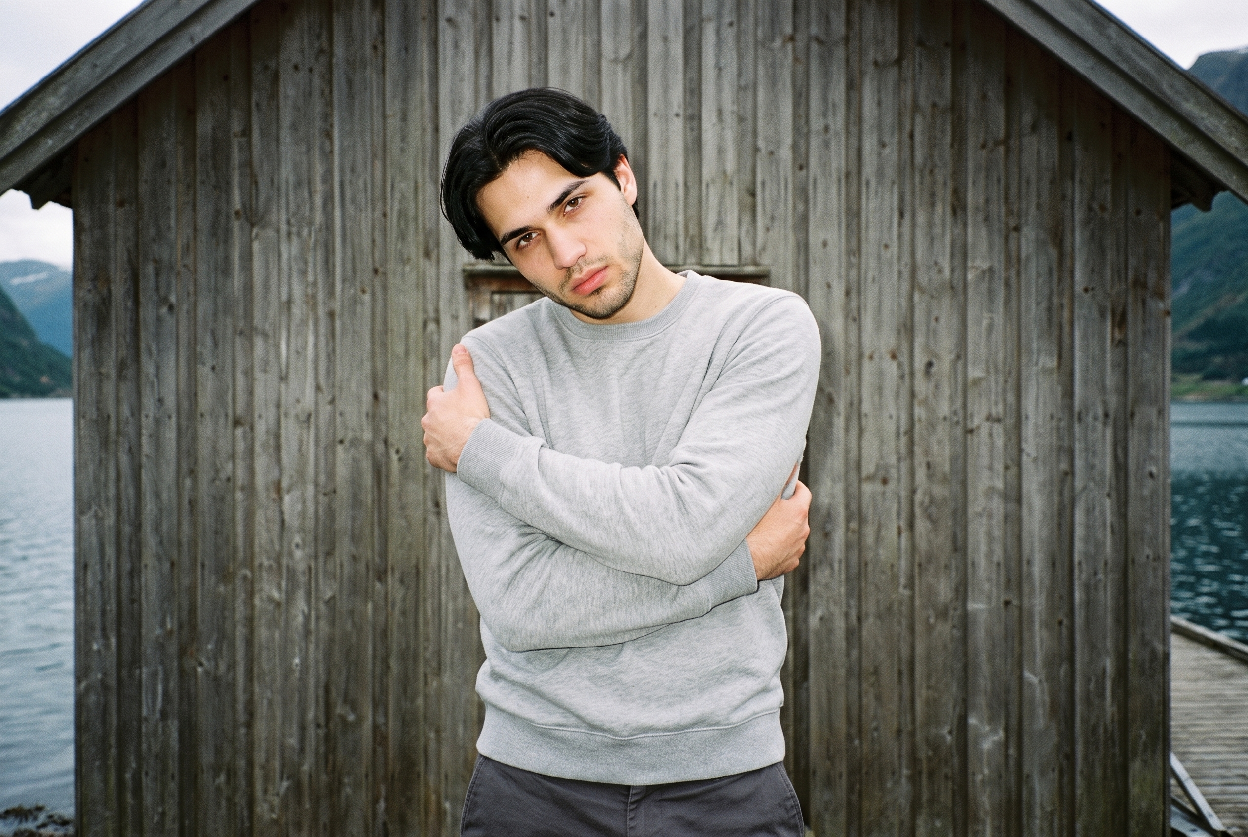 An 18-year-old Slavic male model wears a grey crewneck sweatshirt in an upper-body, front-view composition. He poses with his arms crossed in a self-hug and has a confident facial expression. A Norwegian fjord village appears as the backdrop.
