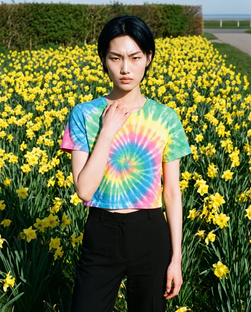 A 23-year-old Korean woman is shown from the upper body in a front view, wearing a tie-dye crop top. One hand rests at her collarbone, and she has a serious expression. A daffodil field forms the backdrop.