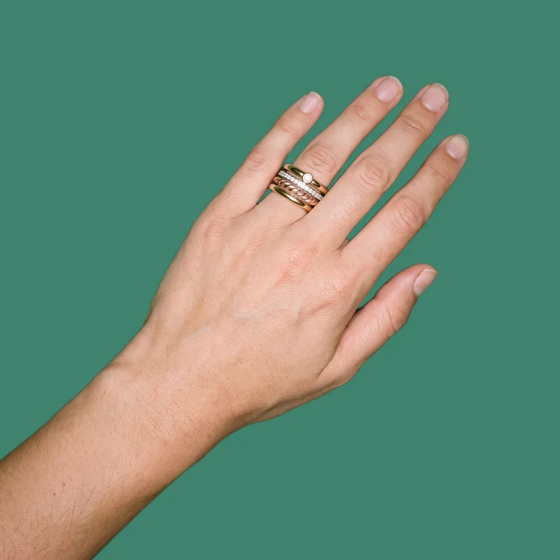 A hand and wrist are shown from above at an angle, wearing a women's mixed-metal stacking ring set. The viridian flat-color backdrop creates a clean, uninterrupted background.