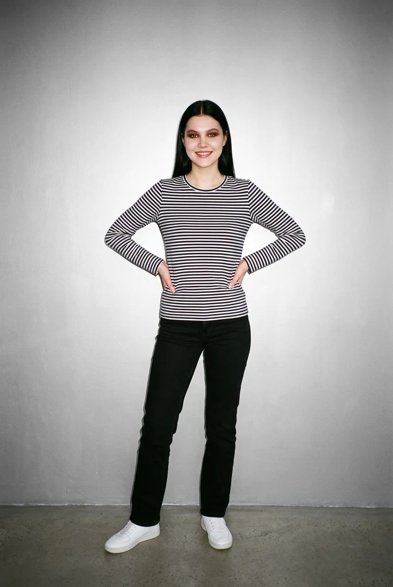 A 19-year-old Caucasian woman wears a women's navy striped long-sleeve tee in a full-body front view. She stands with her hands on her hips and a smiling expression against a Silver Mist studio-wall backdrop.