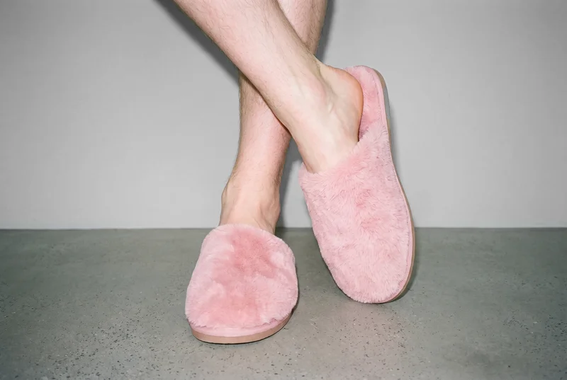 Both feet wear pink fluffy men's slippers in a front three-quarter view, with the ankles crossed. The slippers are shown against a dove grey studio-wall backdrop.
