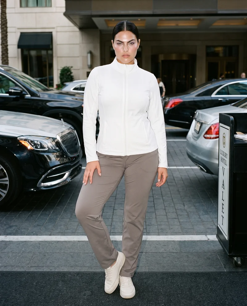 A 23-year-old Caucasian woman wears a white track jacket in a full-body front view. She stands with one hip angled outward and an intense facial expression against a valet stand backdrop.