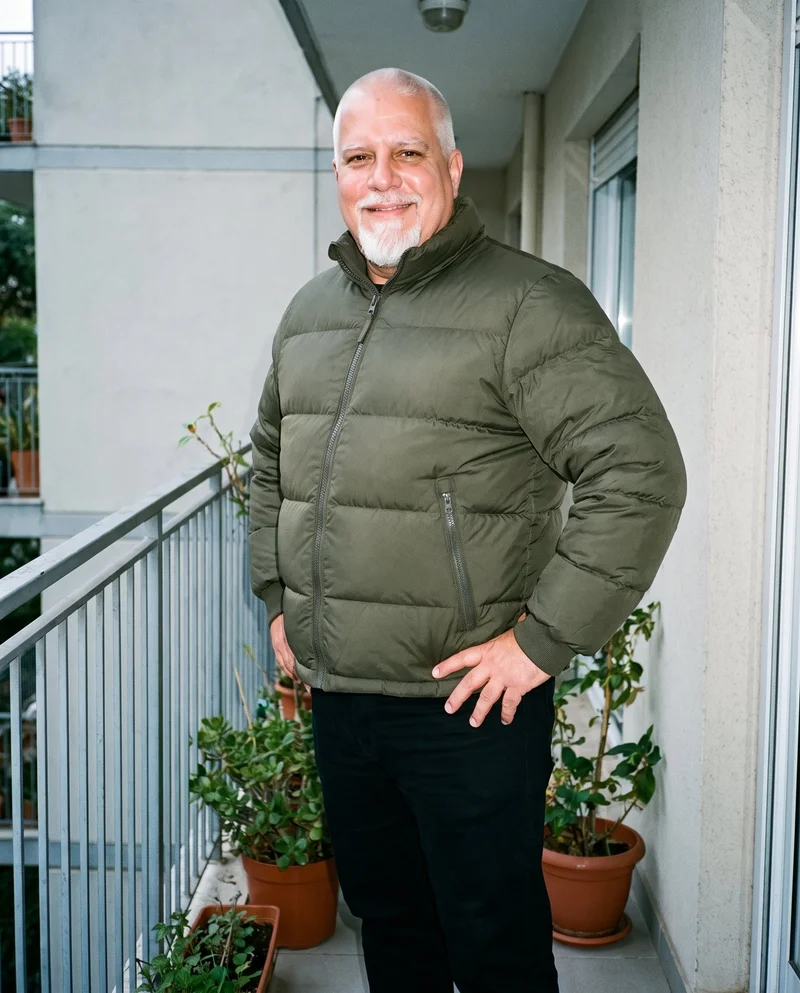 A 56-year-old Caucasian man wears an olive down jacket in a three-quarter-length, angled-front view. He is smiling with one hand on his hip, against a backdrop of balcony plants.