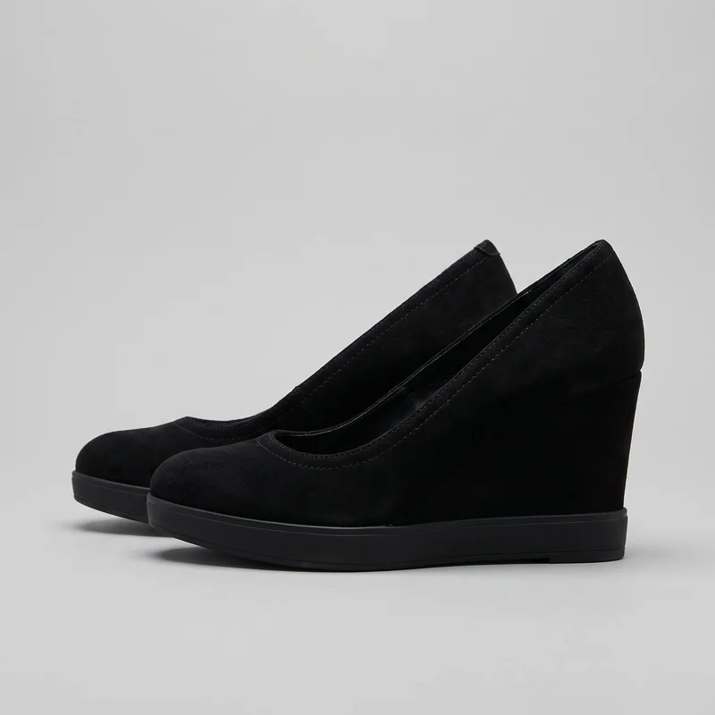 A pair of black women's closed-toe wedge pumps is shown in a side three-quarter view. The shoes have rounded toes, low-cut slip-on openings, closed backs, continuous wedge heels, and platform-like soles with clean thick edges. Their smooth, unembellished uppers feature stitched binding around the toplines and visible seam stitching around the toe openings.