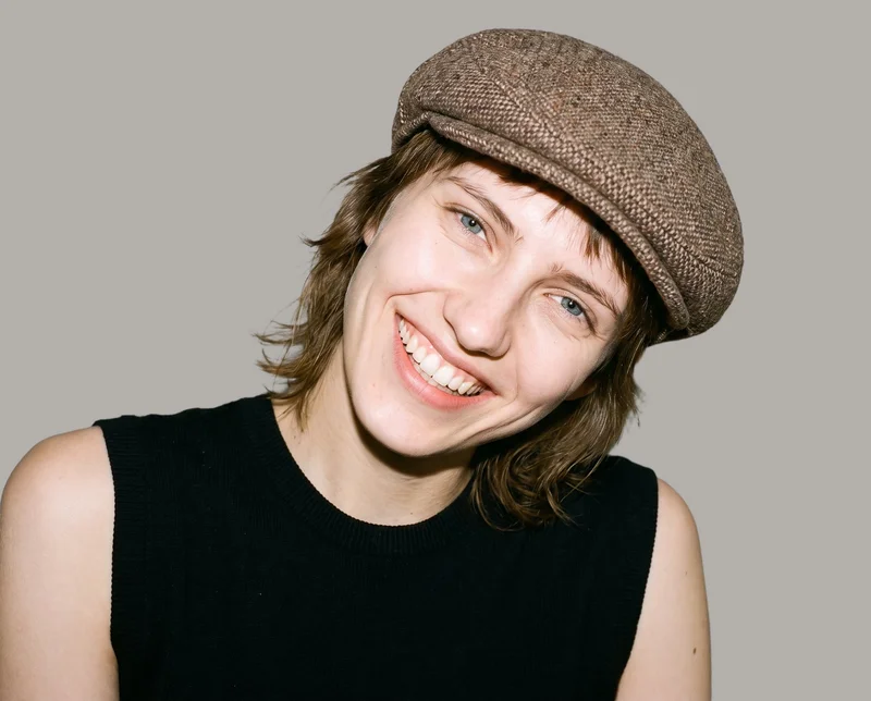 A 20-year-old Caucasian woman wears a brown tweed flat cap in a front-facing bust composition. Her head is tilted and she is grinning against a flat couture grey backdrop.
