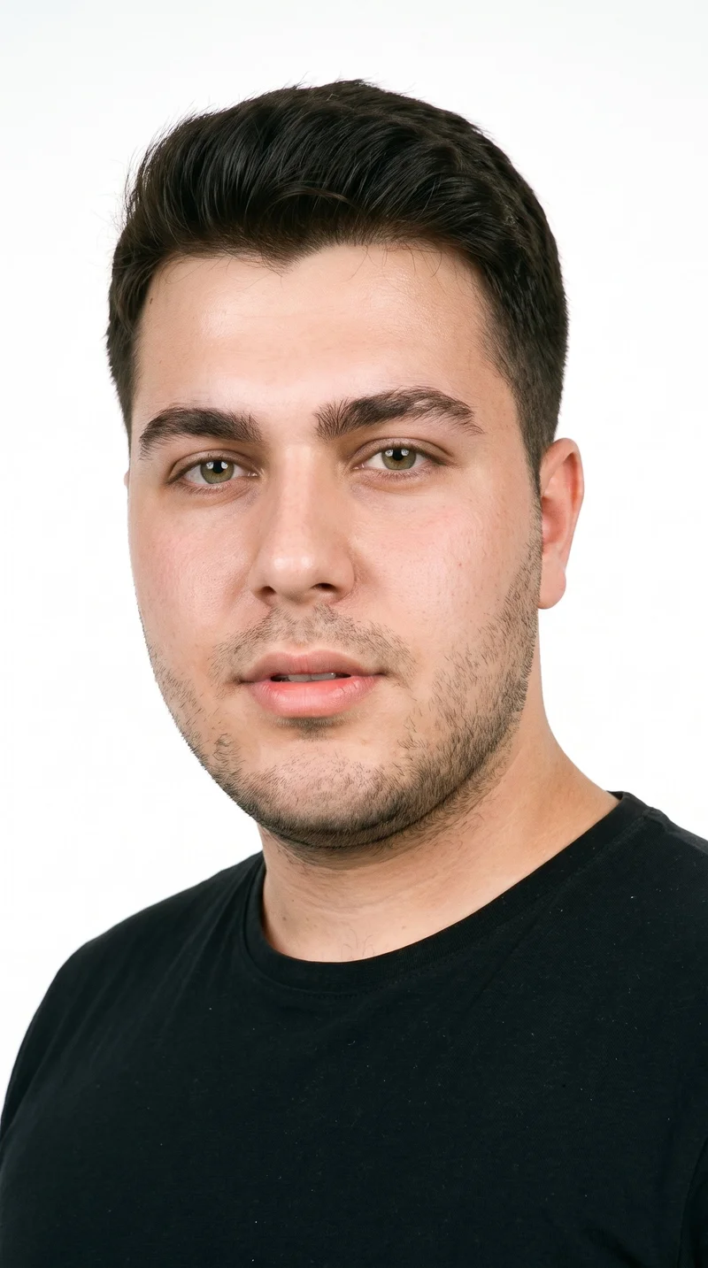 A 20-year-old Turkish man with light-tan skin and an olive-green undertone has a softly structured face with smooth, even features. His eyebrows are thick, and his eyes are green. He has medium lips, dark-brown hair worn in a taper that gradually shortens at the sides and neckline while retaining length on top, and very short facial hair forming a faint, even five-o'clock shadow across the beard area.