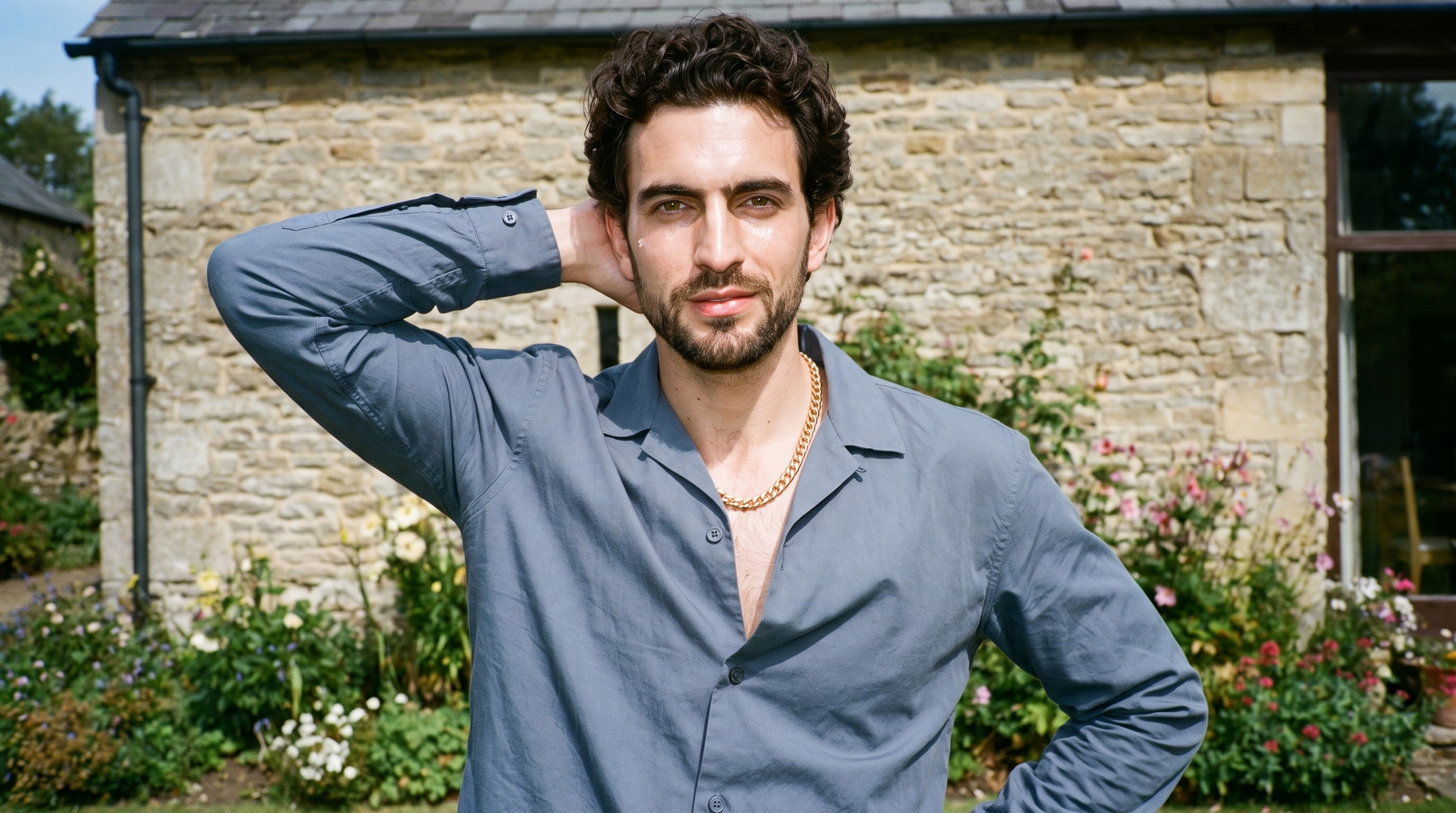 A 33-year-old Caucasian man is shown from the front in an upper-body view wearing a gold chain necklace, with one hand behind his neck and a confident expression. A cottage garden forms the backdrop.