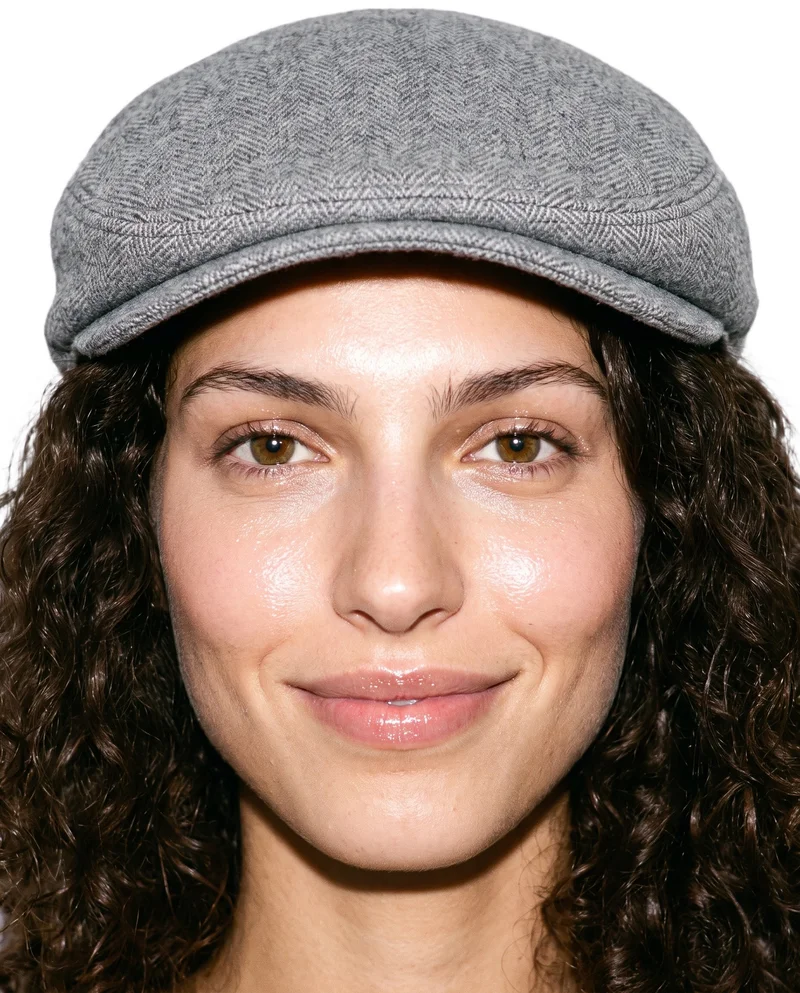 A smiling 22-year-old Caucasian woman wears a women's grey wool flat cap. She is shown from the head up in a front view with her head level, against a pure white backdrop.