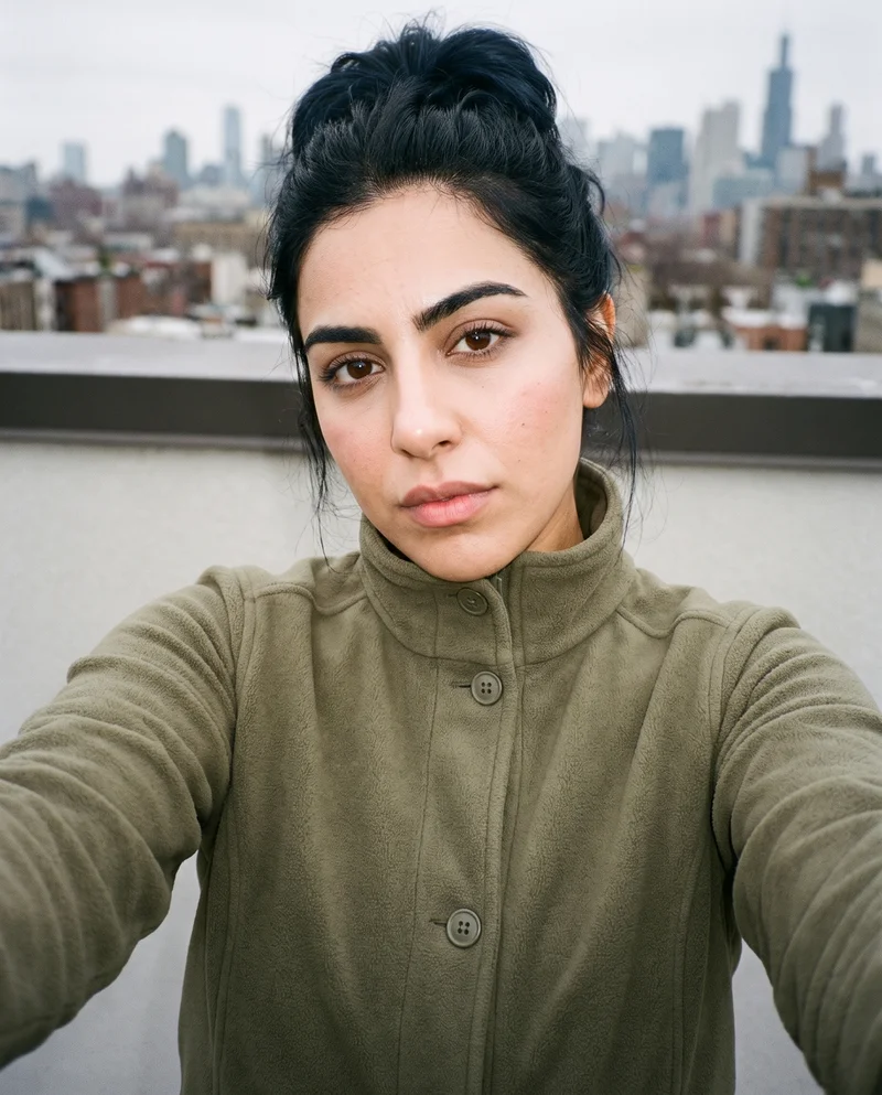 A 23-year-old Arab woman wears a women's olive fleece jacket in a front-facing selfie with her head tilted and a serious expression. A rooftop cityscape forms the backdrop.