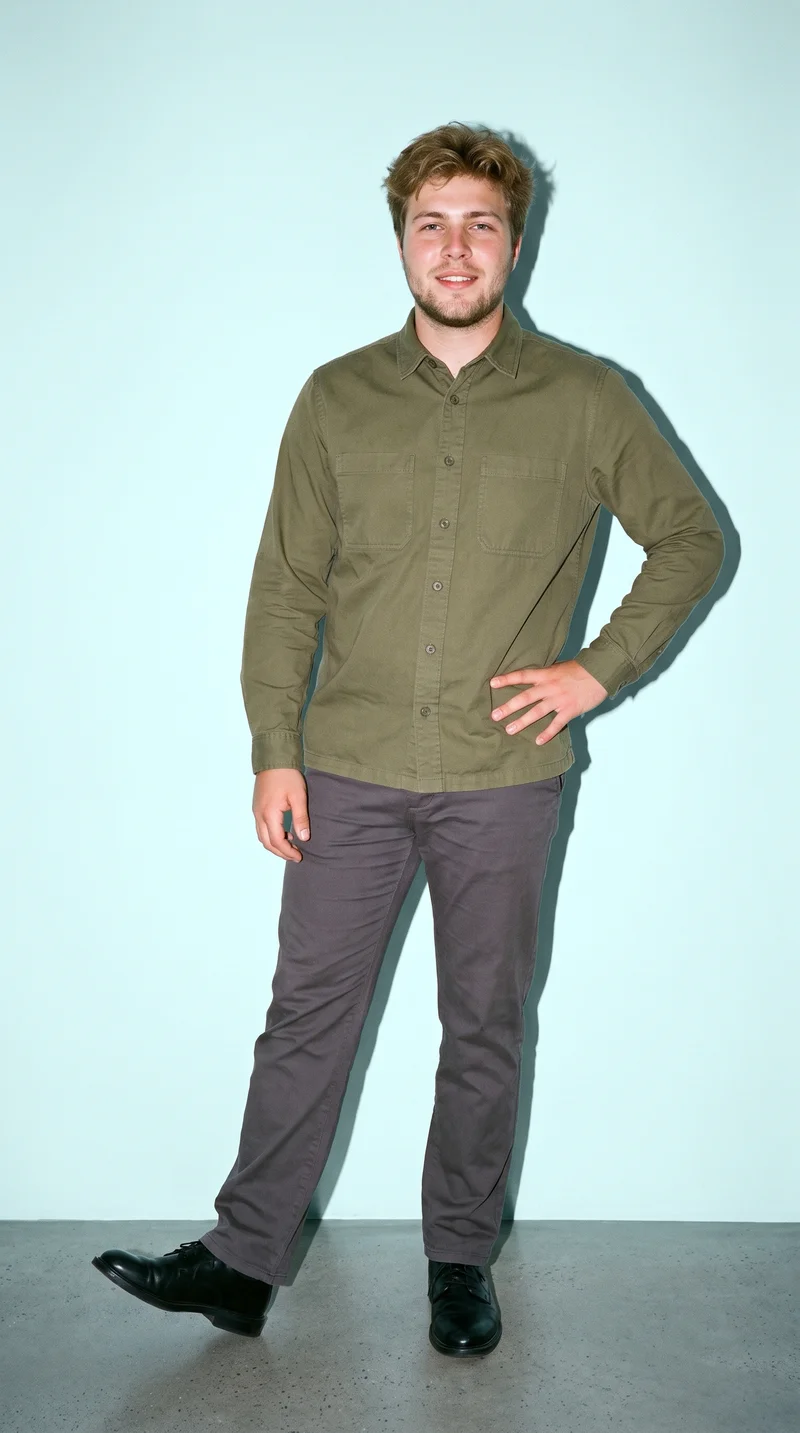 An 18-year-old Caucasian man wears an olive cotton overshirt in a full-body front view. He is smiling with one foot forward against a light cyan studio-wall backdrop.