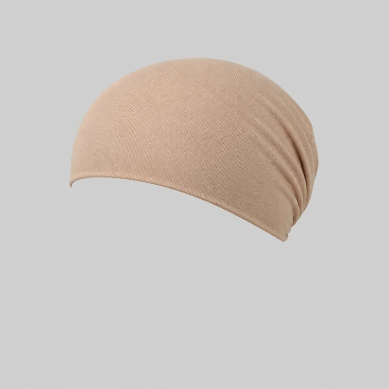 A beige cotton underscarf is shown as a standalone product, with a rounded close-fitting crown, a curved lower edge, and gathered ruching folds tapering toward one rear side. Fine ribbed texture is visible across the mostly complete silhouette. No closures, straps, brim, ties, logos, prints, patches, embroidery, or embellishments are visible.