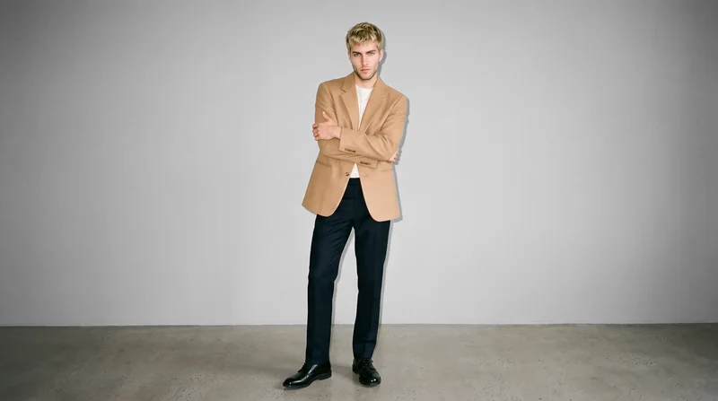 A 23-year-old Caucasian man wears a camel oversized blazer in a front-view environmental composition. He stands in a self-hug pose with a neutral expression against a dove grey studio-wall backdrop.