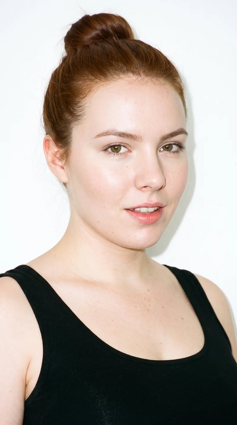 This 18-year-old Caucasian woman has very pale skin with a peach undertone and a heart-shaped face, with a wider forehead narrowing to a pointed chin. She has thin eyebrows, blue eyes, and full lips. Her warm reddish-brown hair is wound into a rounded bun at the back of her head.