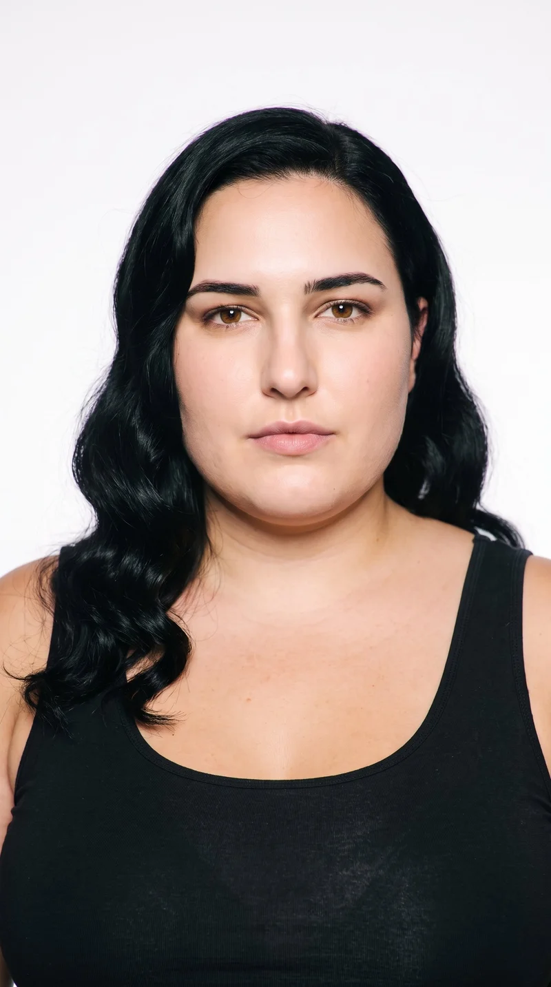 This 26-year-old Caucasian woman has lightly tanned fair skin with a warm golden undertone. Her square face has a strong, defined jaw and a broad forehead, creating an angular, structured appearance. She has medium-thickness eyebrows, brown eyes, and thin lips. Her black hair is styled in large, polished waves with a smooth, glamorous finish.