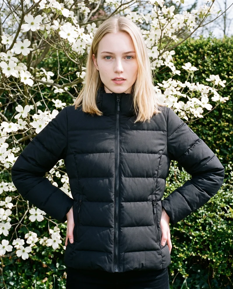 A 19-year-old Caucasian woman is shown from the front in an upper-body view wearing a black down jacket. She stands with her hands on her hips against a dogwood tree grove backdrop, with an editorial facial expression.